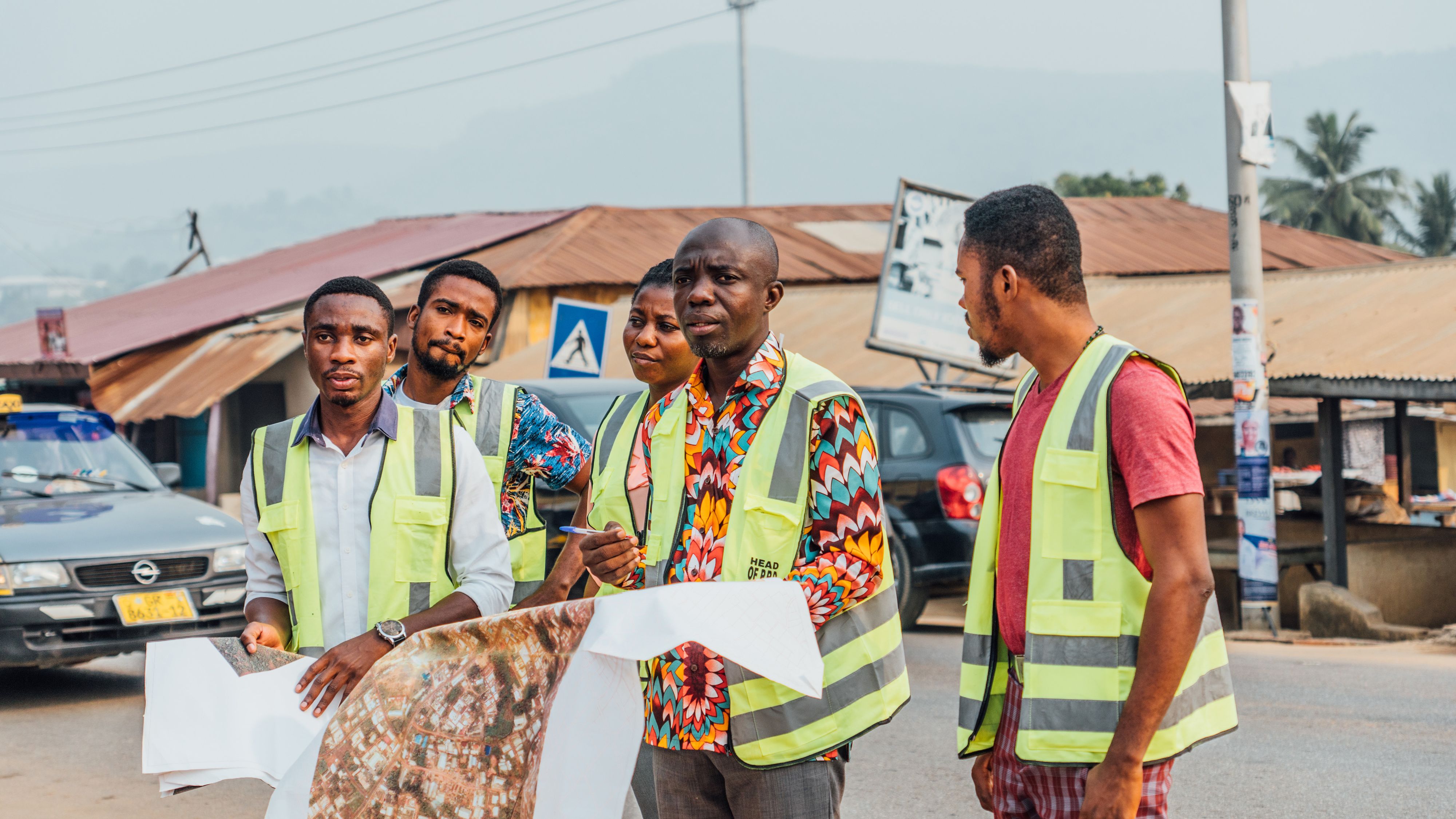 Five people in reflective vests stand on a street in Ghana, discussing a large map. Vehicles and buildings are in the background. 