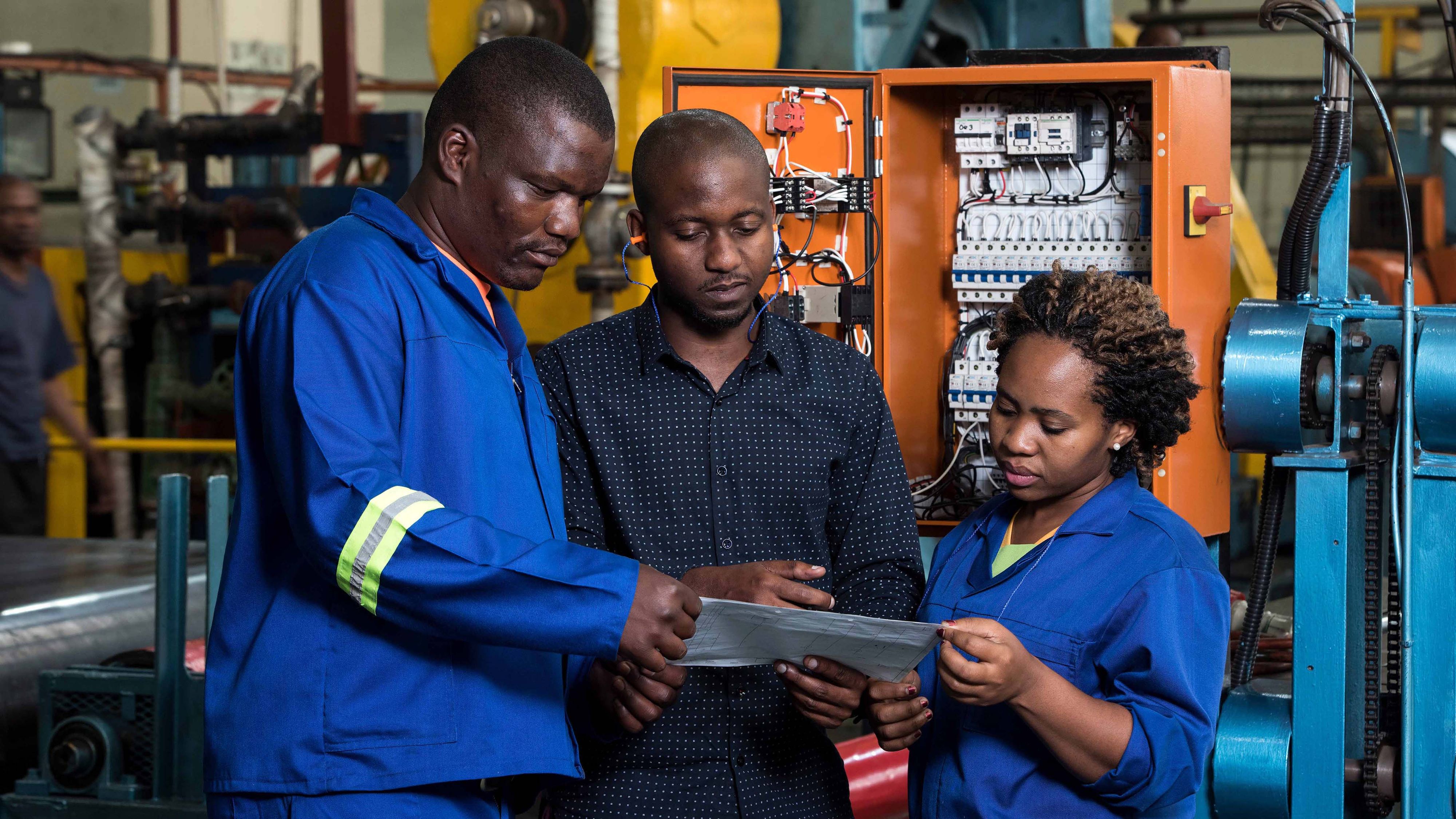 Three professionals in a factory reviewing a document in front of industrial equipment. 
