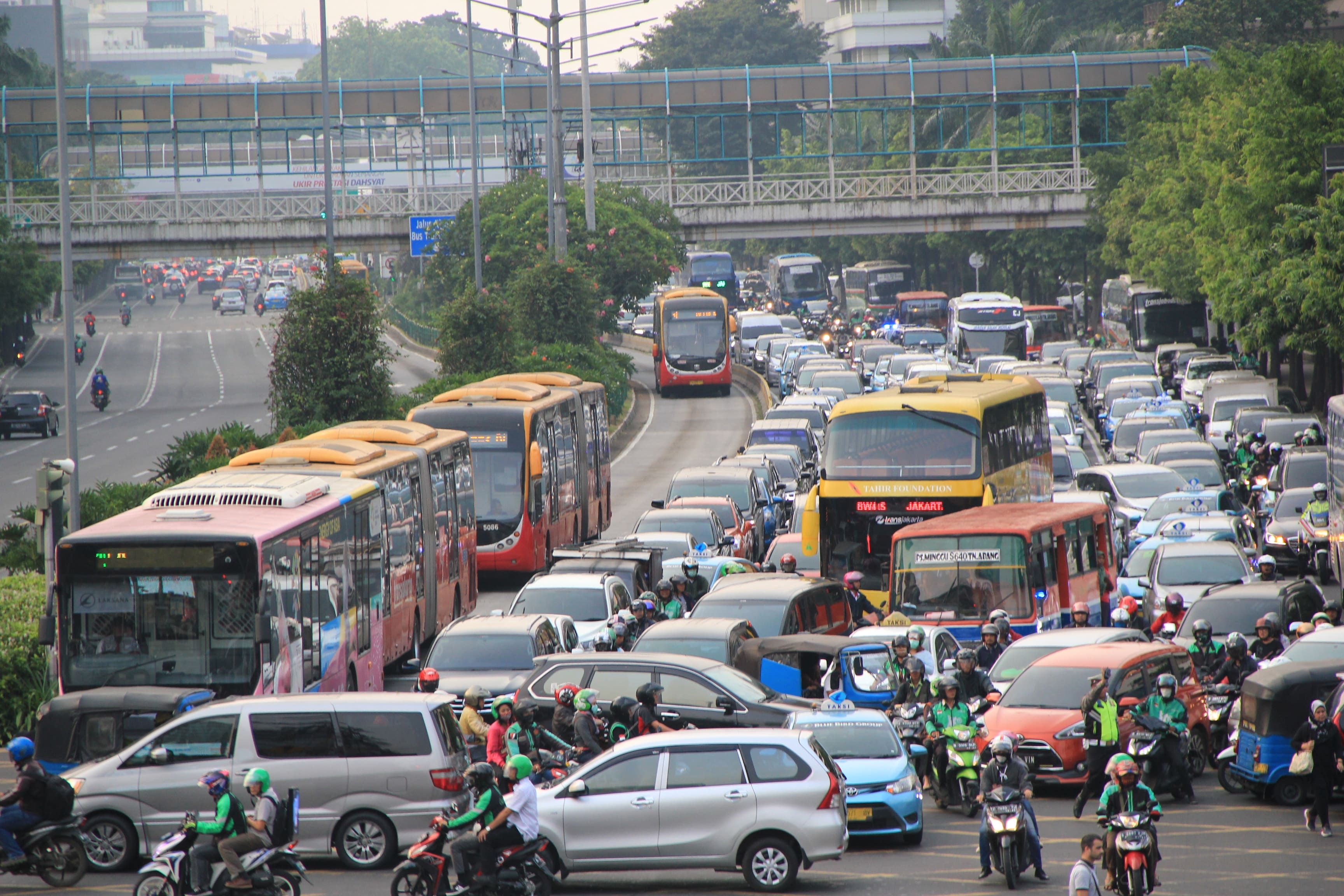 Une rue bondée à Jakarta avec un trafic dense, incluant des bus, des voitures et des motos. Un pont piétonnier et des espaces verts sont visibles à l'arrière-plan.